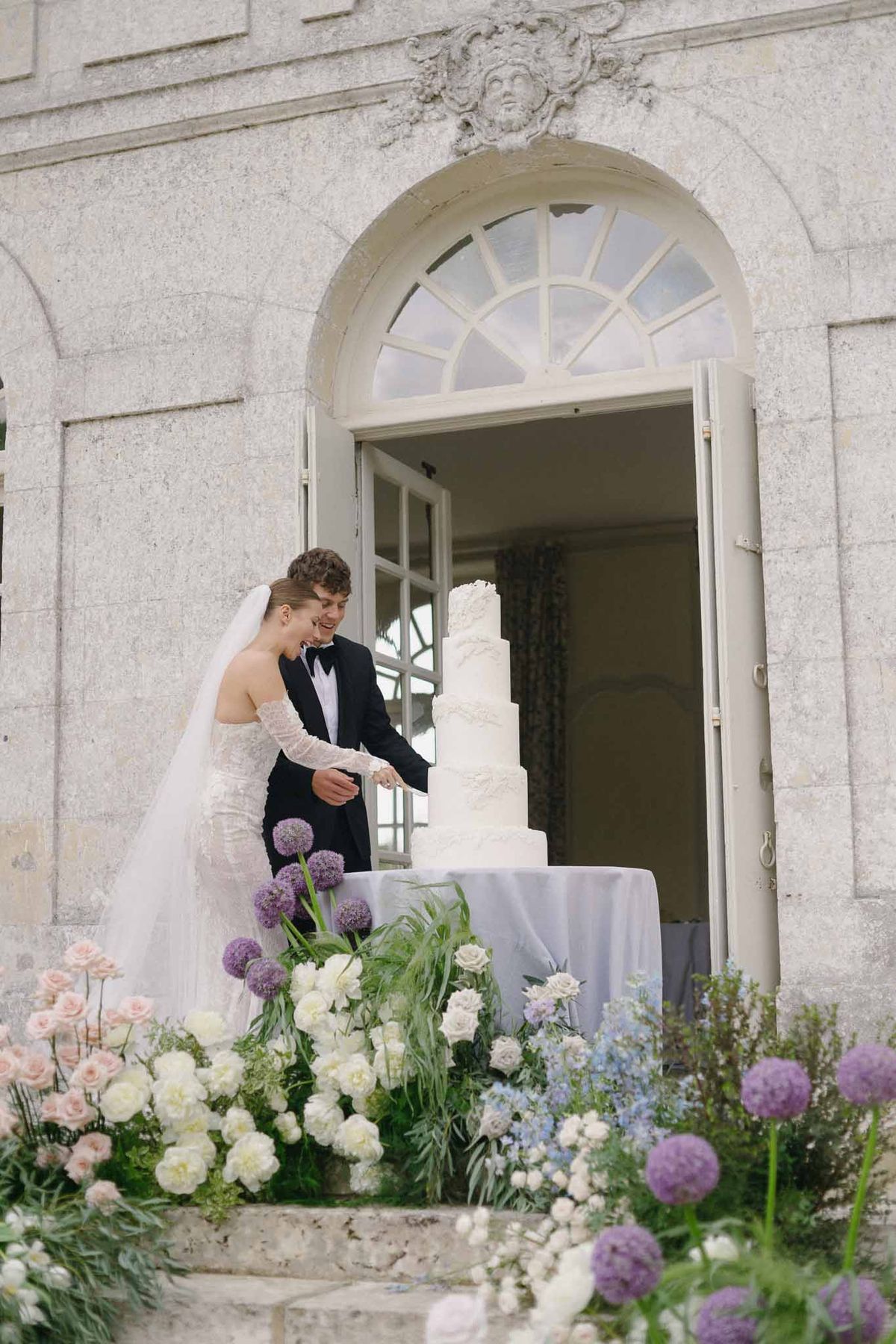 Bride and groom cutting wedding cake outdoors at classical stone venue with floral arrangements