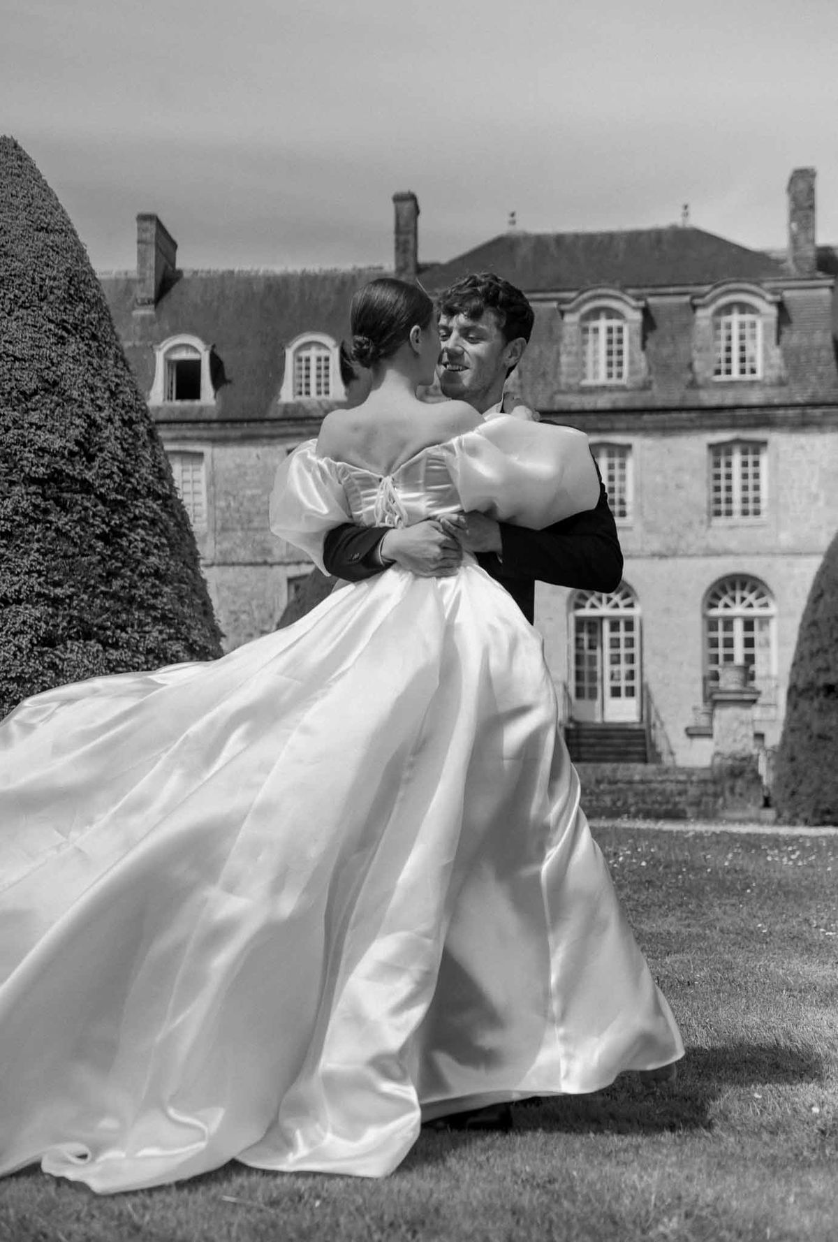 Groom lifting bride in white gown at classical manor house with topiary hedge