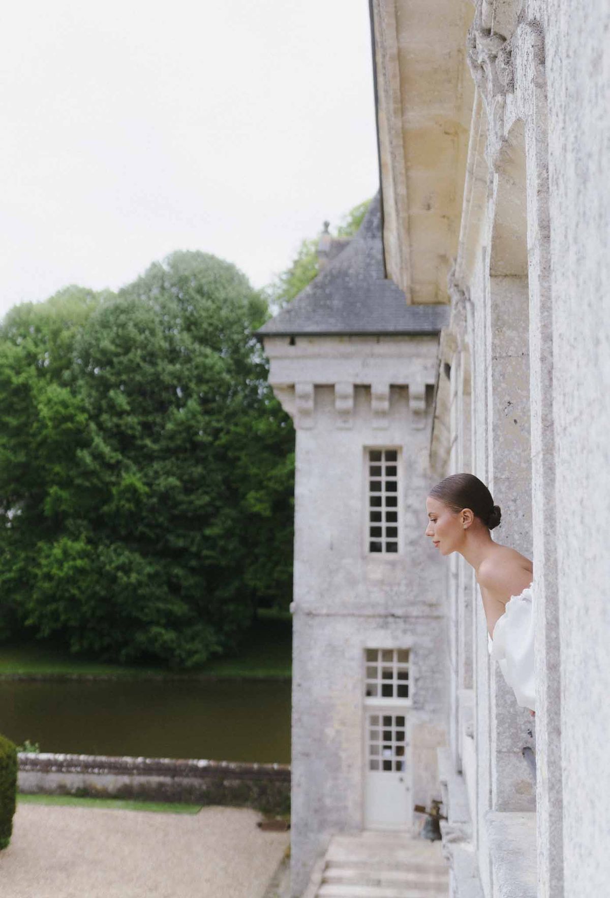 Bride in white gown peeking around stone château corner in courtyard