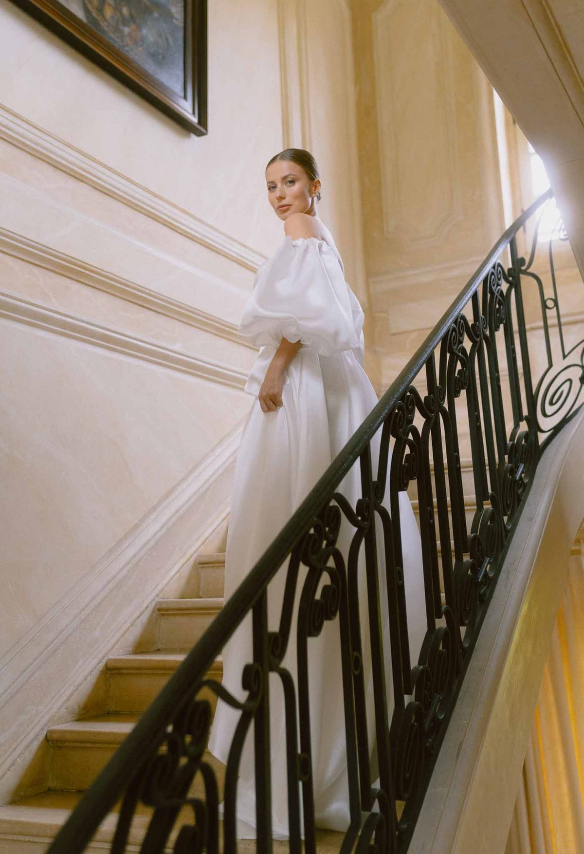 Bride in white gown descending grand staircase in classical European interior with ornate molding