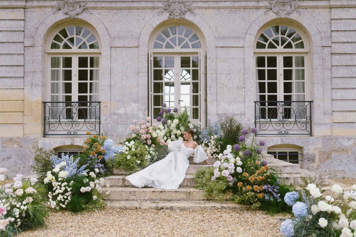 Bride in white gown sitting on stone steps of classical château with garden florals