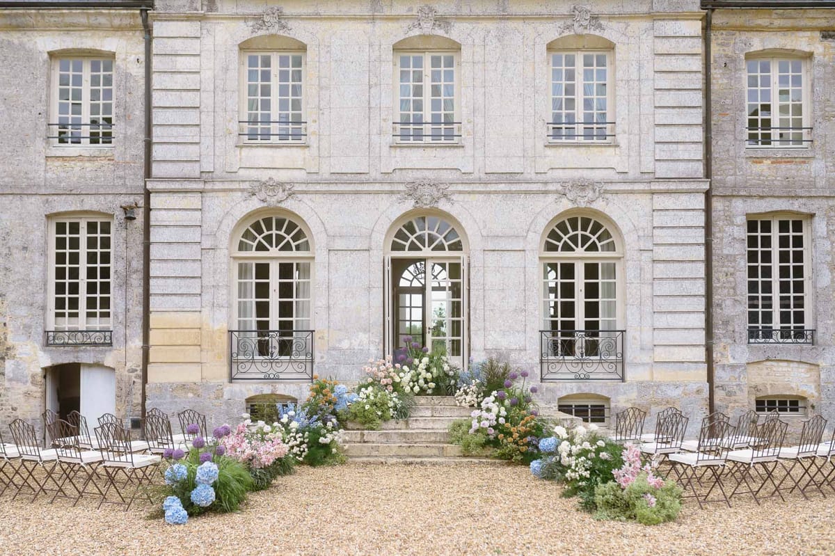 Garden-style ceremony with blue hydrangea and pink floral aisle borders before limestone chateau staircase