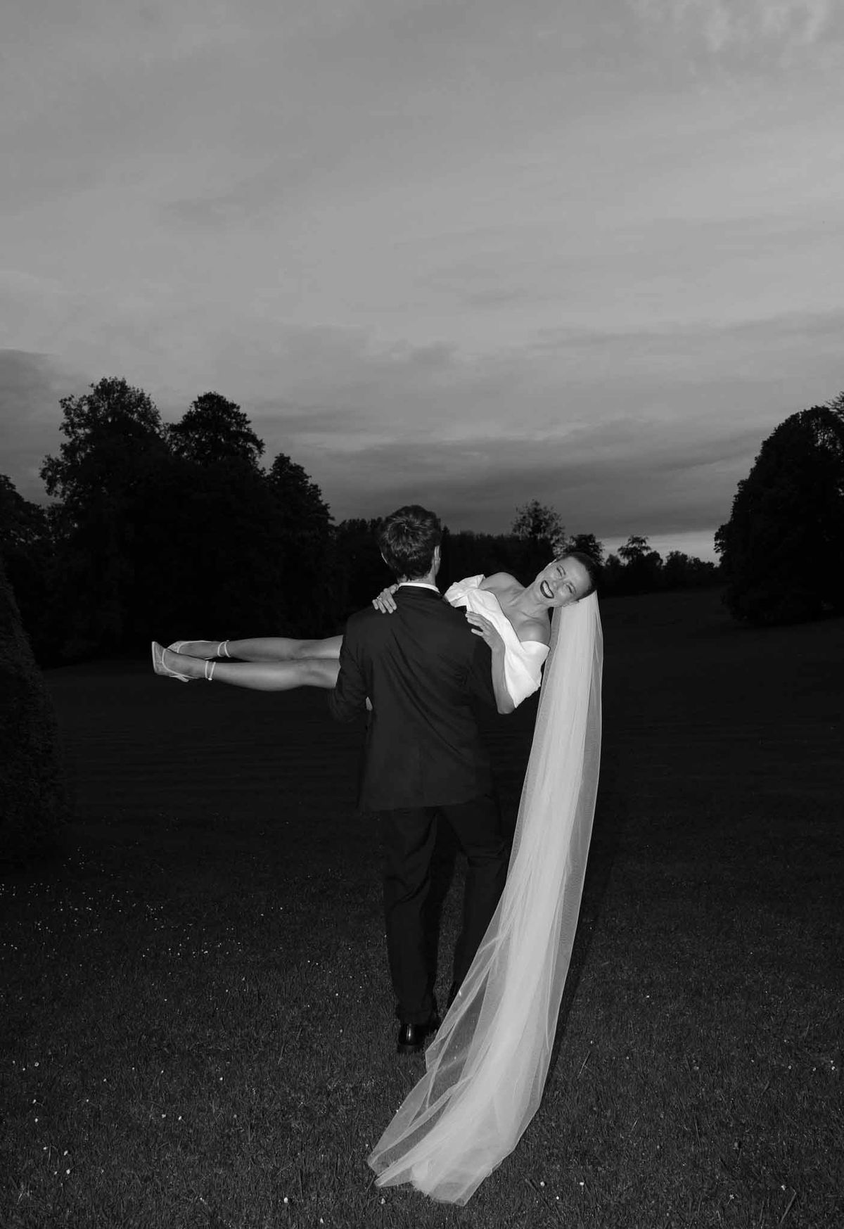 Groom carrying bride across field in black and white portrait at countryside estate