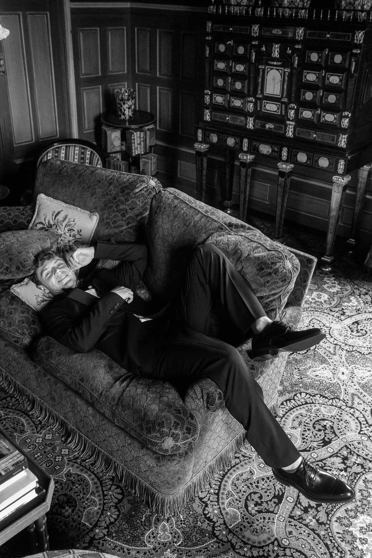 Groom in formal suit relaxing on decorative sofa in elegant getting ready lounge