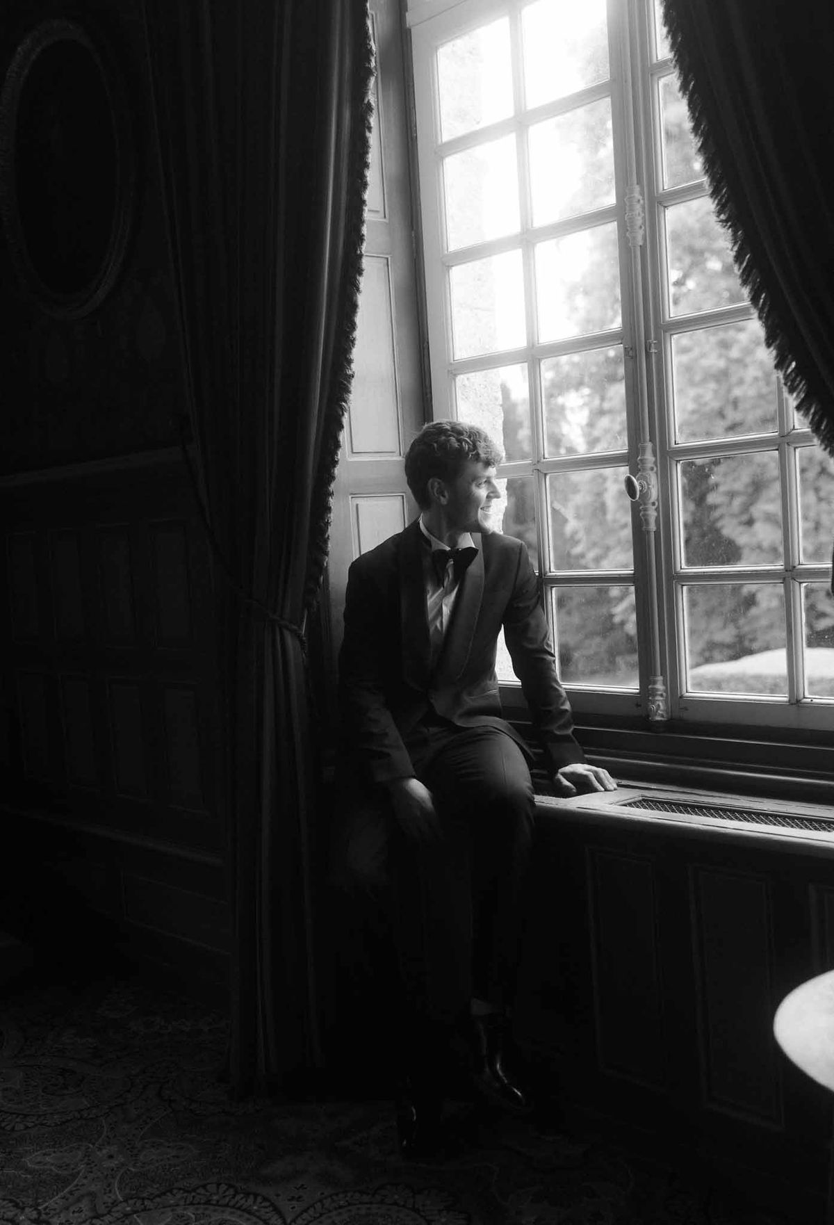Groom in black tuxedo sitting contemplatively at window before wedding ceremony in historic interior