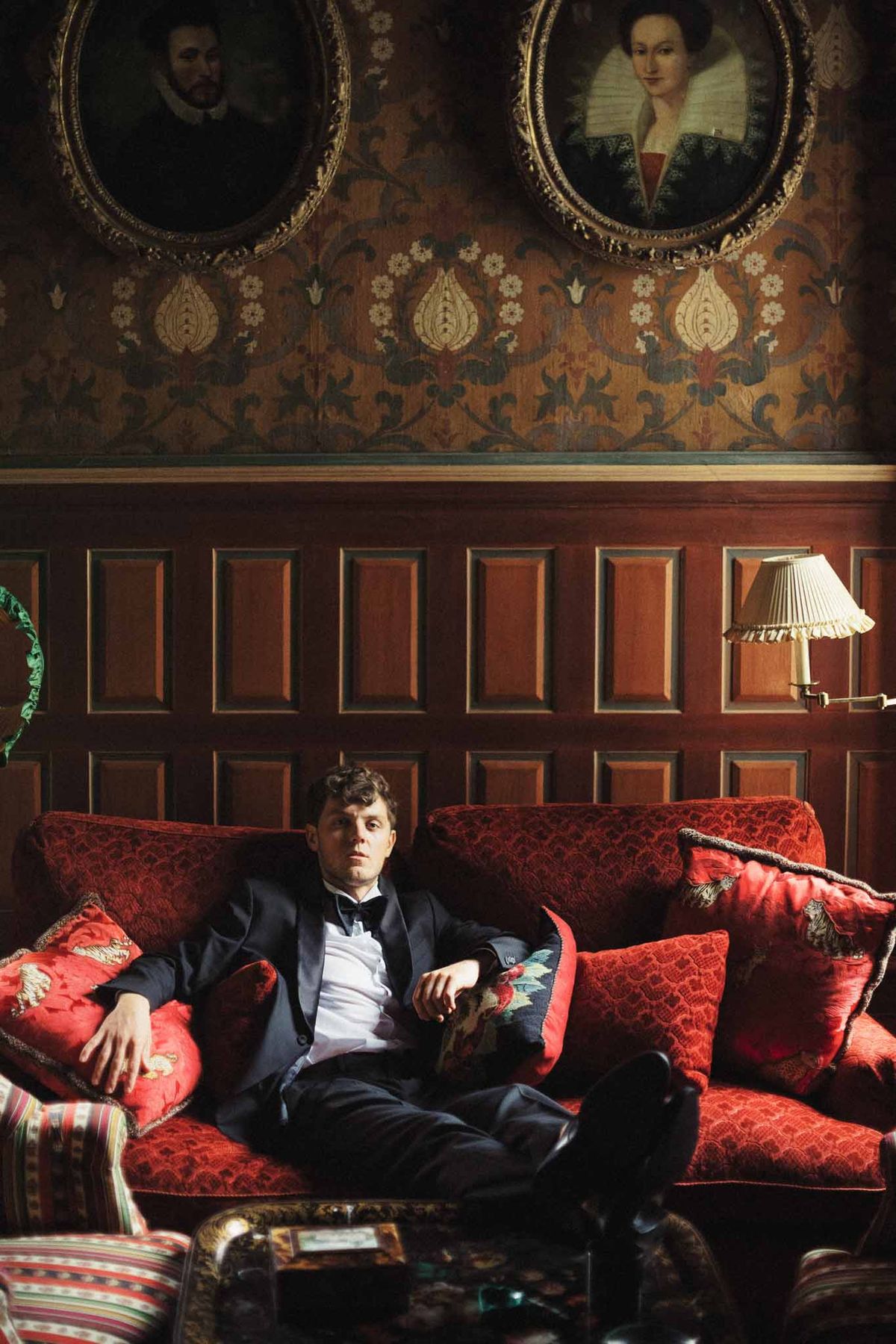 Groom in navy tuxedo and bow tie sitting on red damask sofa in ornate classical interior room