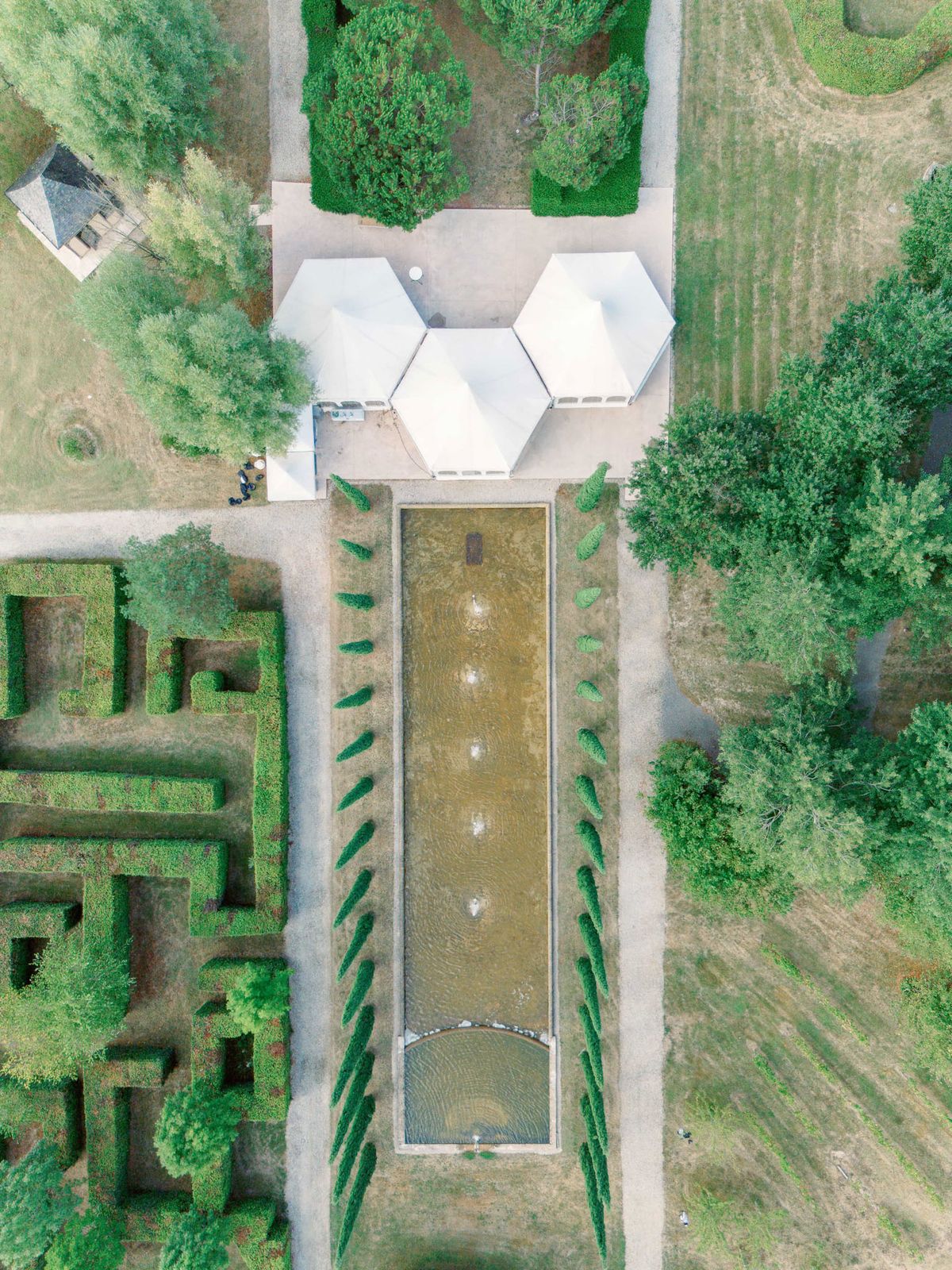 Aerial estate with reflecting pool, conical topiary rows, geometric hedge maze, and white hexagonal event tents