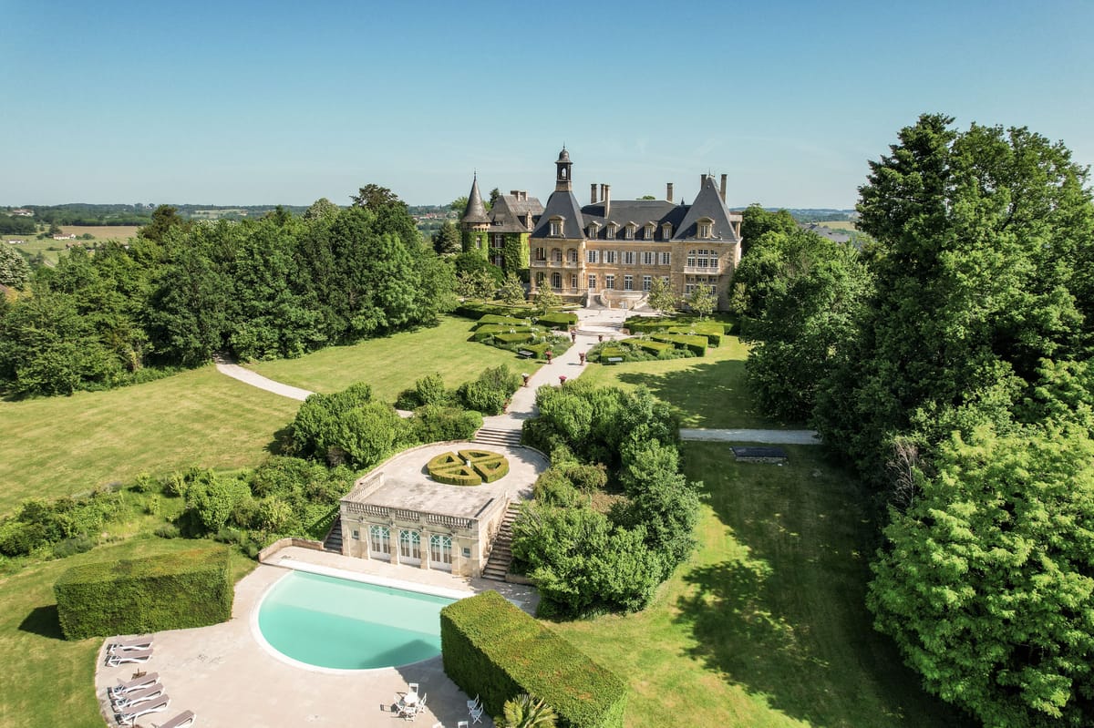Aerial golden limestone chateau with bell tower, curved pool, box parterre, and orangery pool house