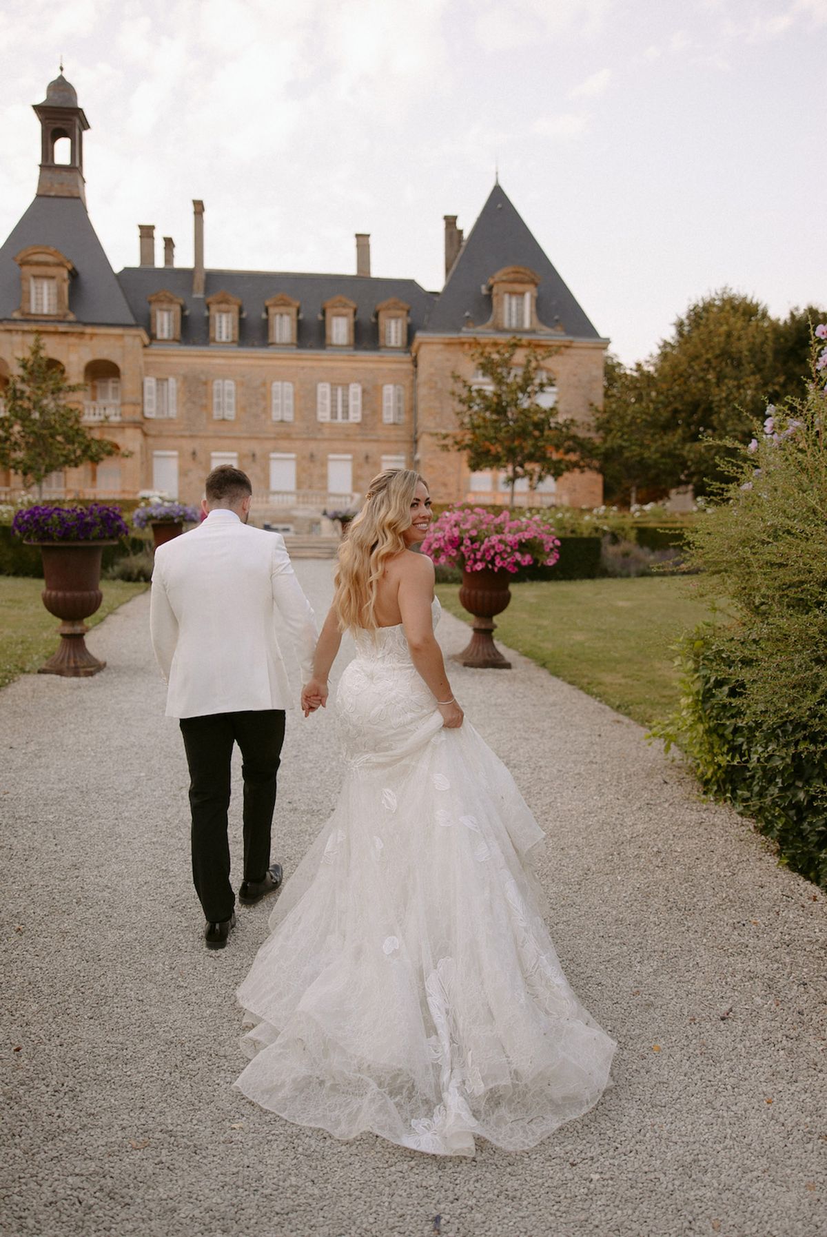 Couple from behind walking gravel path toward slate-turret chateau with terracotta urns and trailing bridal train