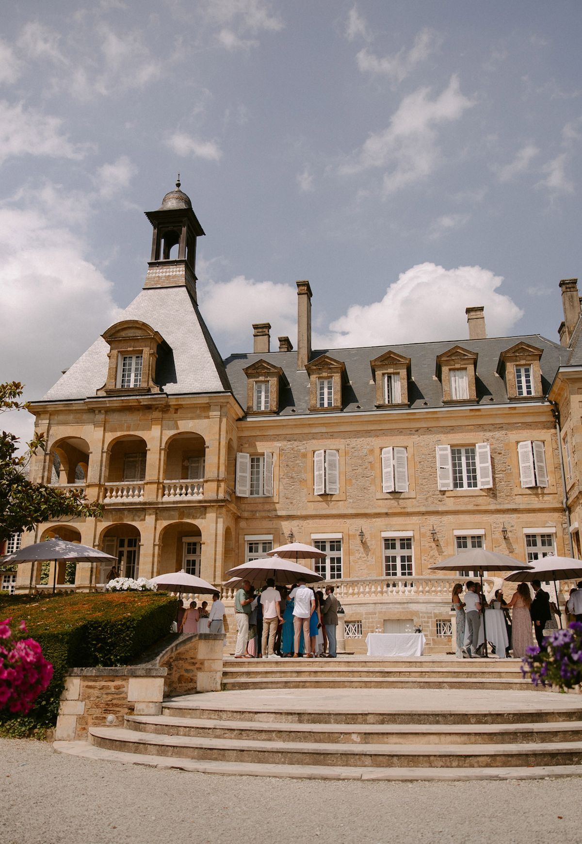 Thirty guests under mauve umbrellas on limestone chateau terrace with mansard roof and corner spire
