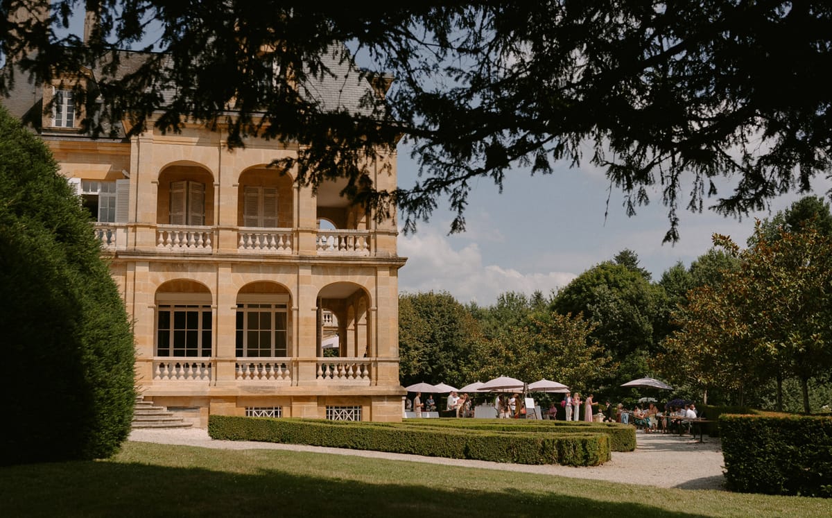 25 guests under taupe parasols on gravel terrace beside honey limestone chateau with arched arcades