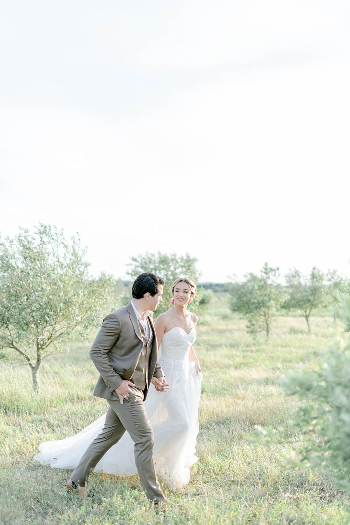 Bride in strapless ivory gown and groom in taupe suit walking hand in hand through olive tree landscape
