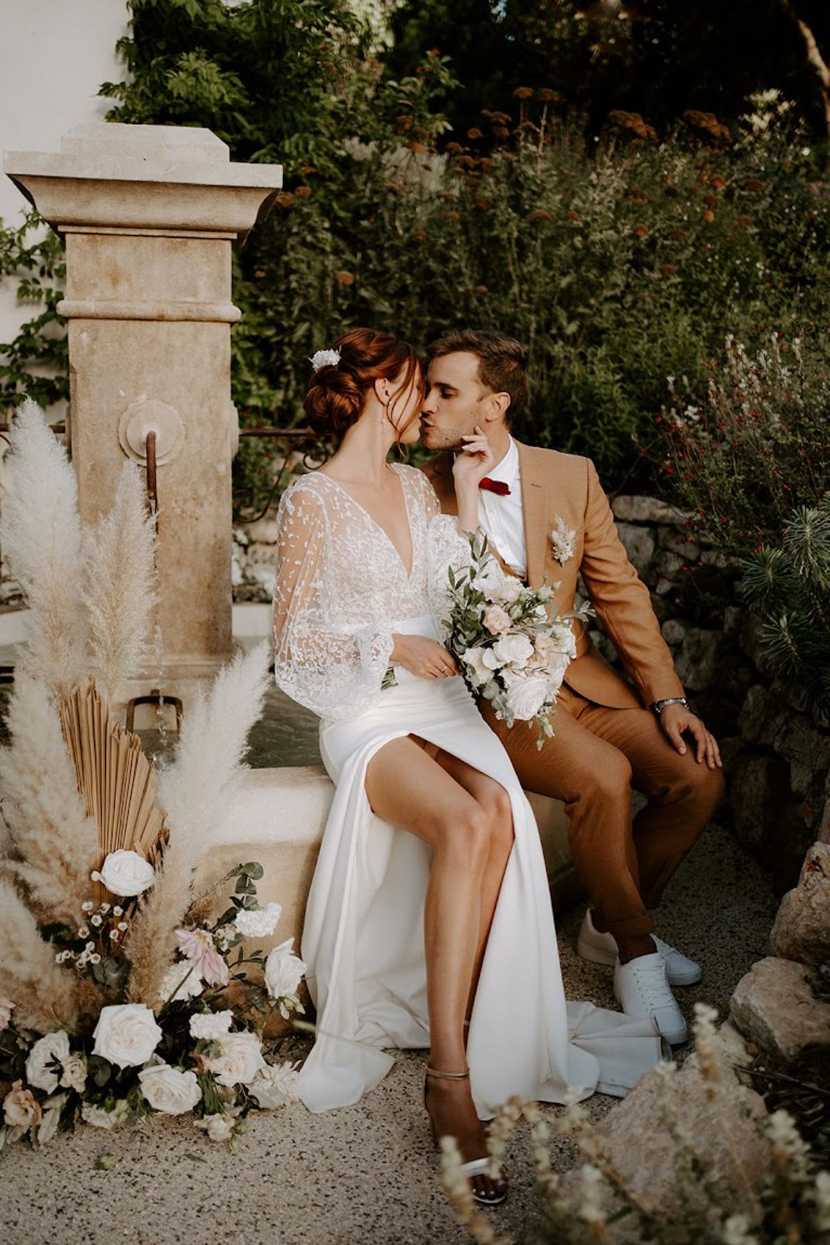 Bride and groom kiss on stone fountain steps surrounded by pampas grass and blush rose arrangements in garden