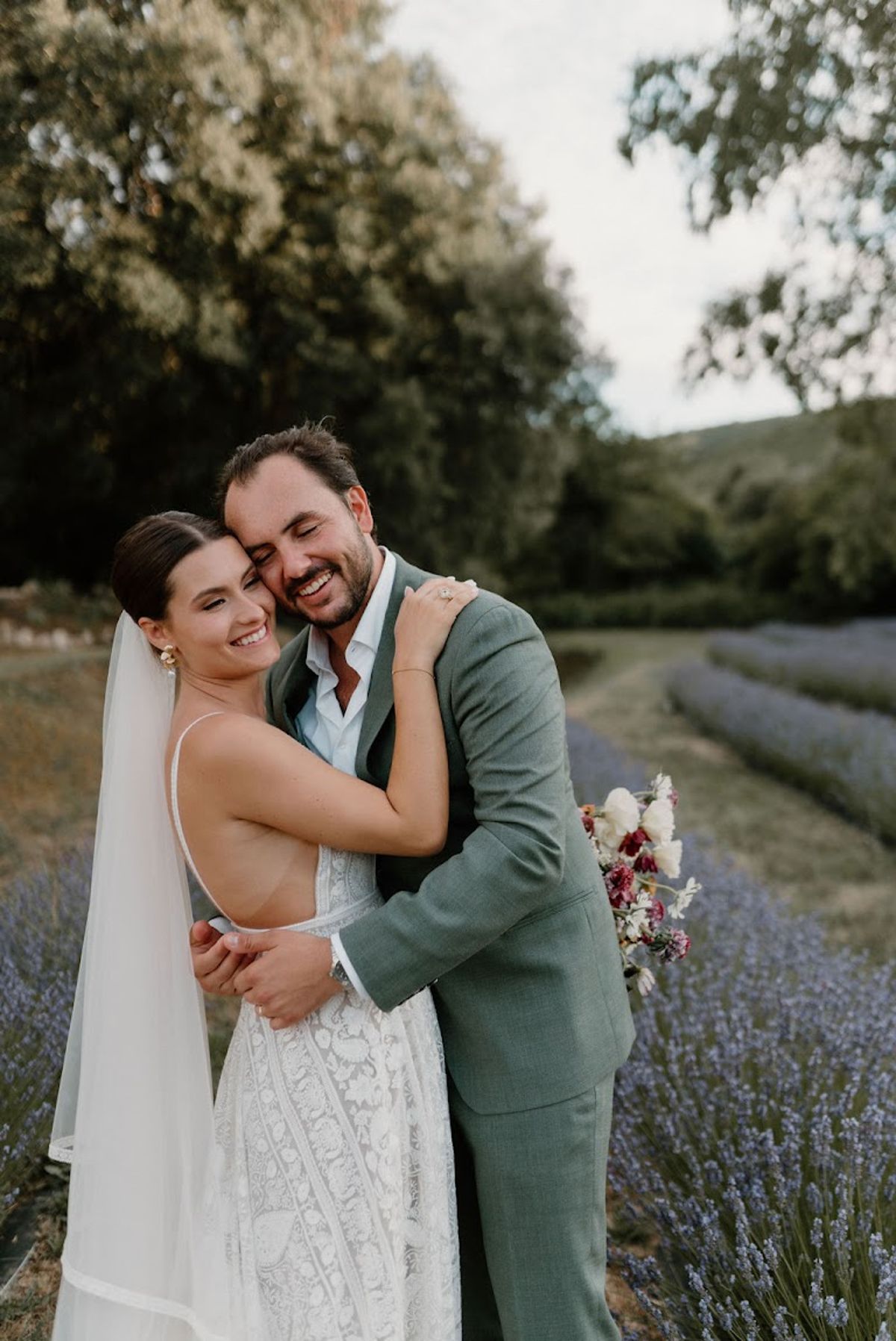 Bride and groom embracing in lavender field with burgundy and cream ranunculus bouquet at golden hour