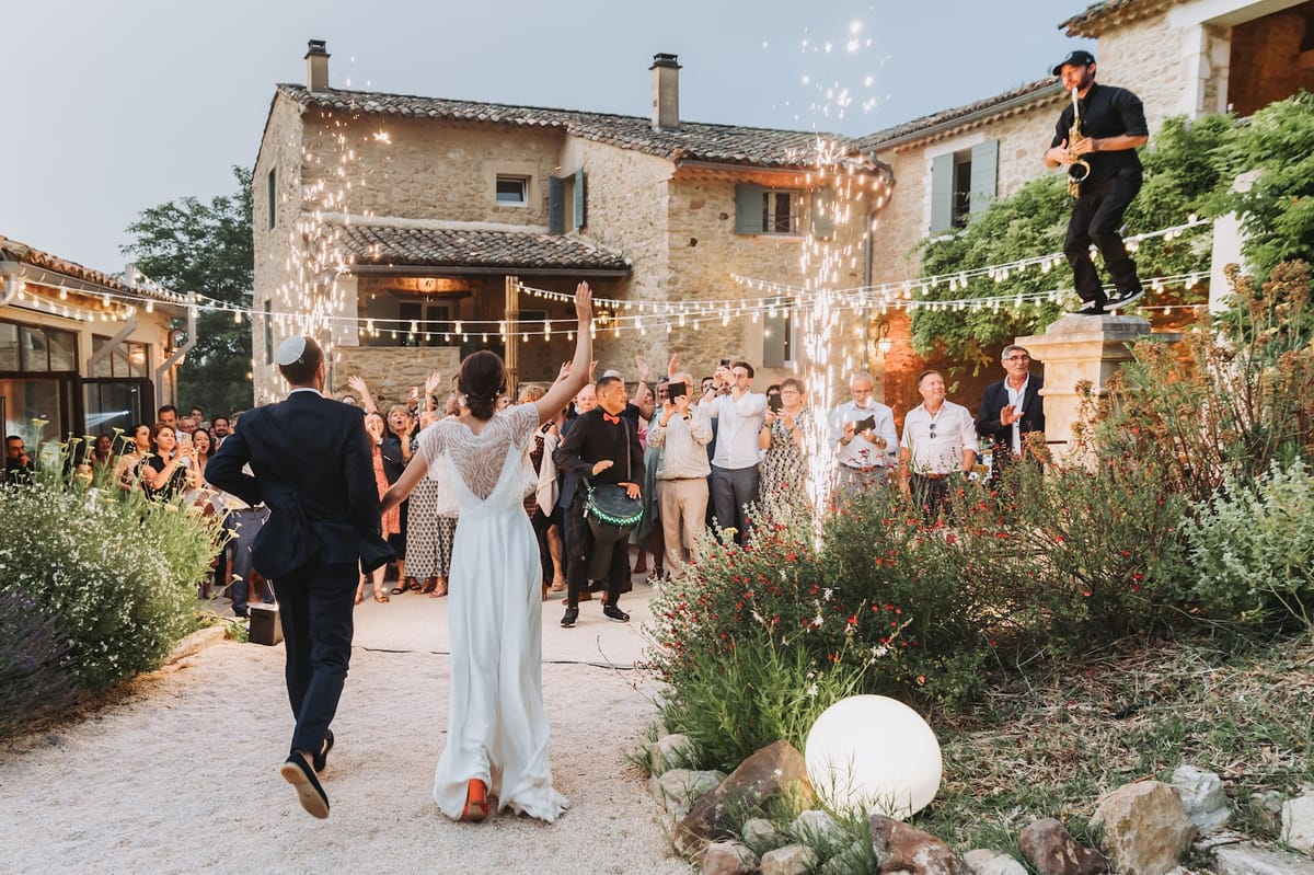 Couple entering Provencal courtyard reception with sparkler fountains, saxophonist, and cheering guests