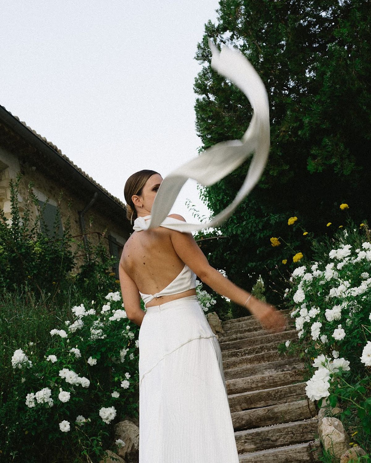 Bride from behind in two-piece white halter crop top and skirt with billowing veil on rustic steps beside white roses