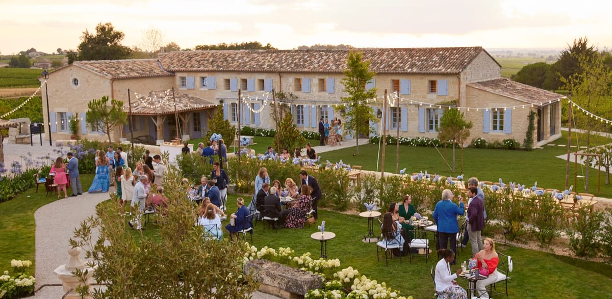 Fifty guests at bistro tables on lawn with festoon lights before stone farmhouse with vineyard beyond