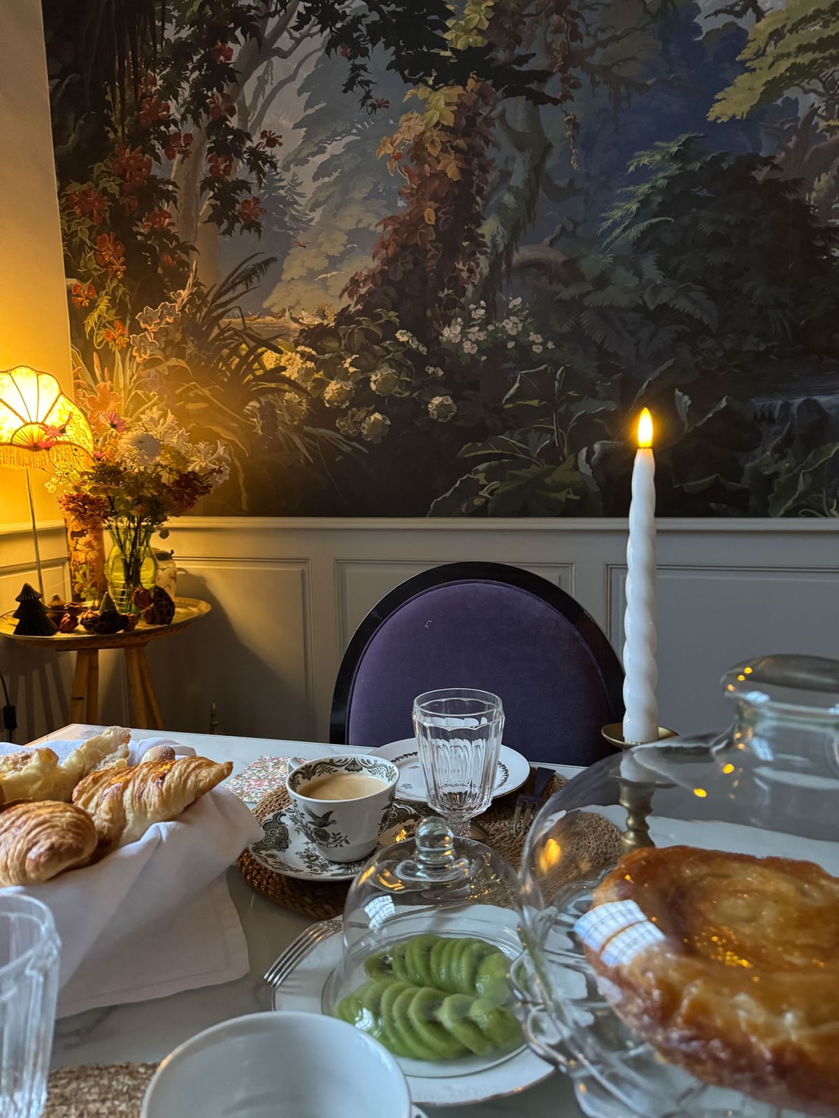 Close-up of a wedding morning breakfast table with croissants, coffee, kiwi, and a lit candle against botanical mural wall...