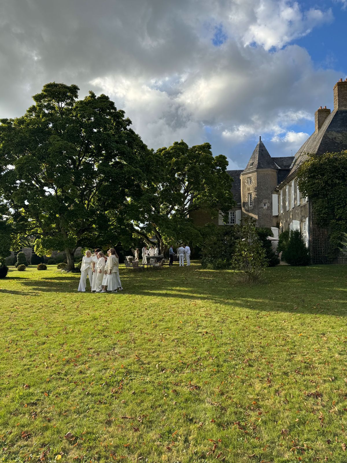 15 guests in all-white dress code gathered on manor lawn with stone tower and bistro drinks station