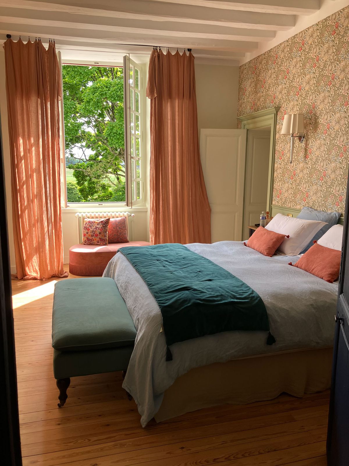 Chateau bedroom with botanical wallpaper, teal velvet throw, salmon curtains, sage armoire, and exposed white beams