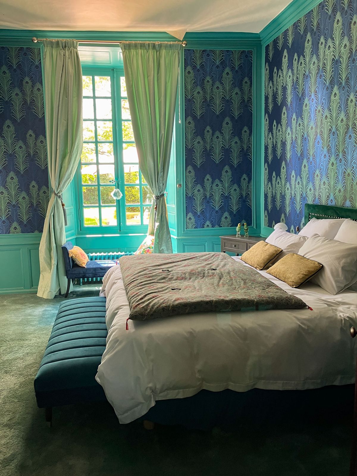 Chateau bedroom with navy peacock wallpaper, turquoise paneling, green velvet headboard, and French windows