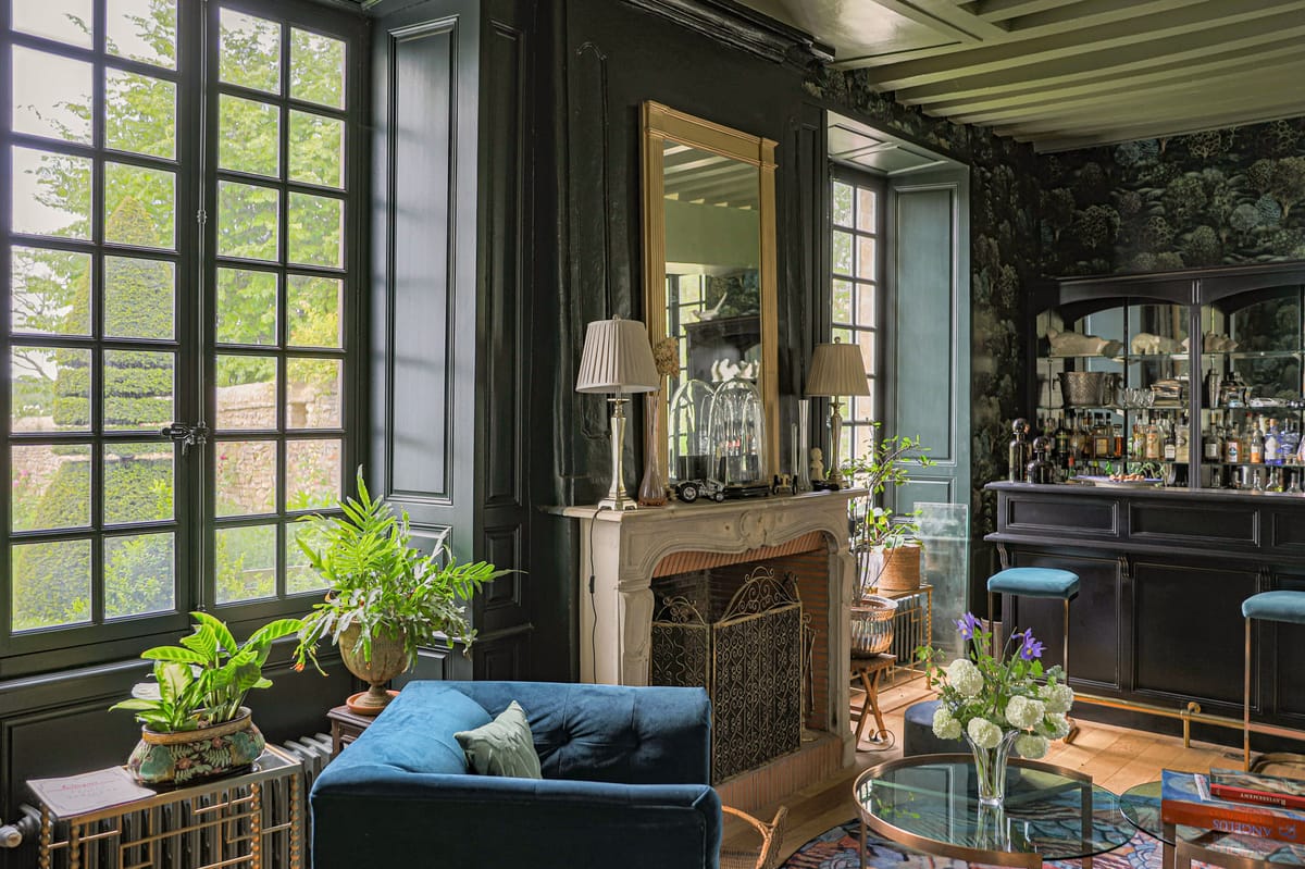 Chateau salon with forest green paneled walls, white marble fireplace, teal bar cabinet, and French windows