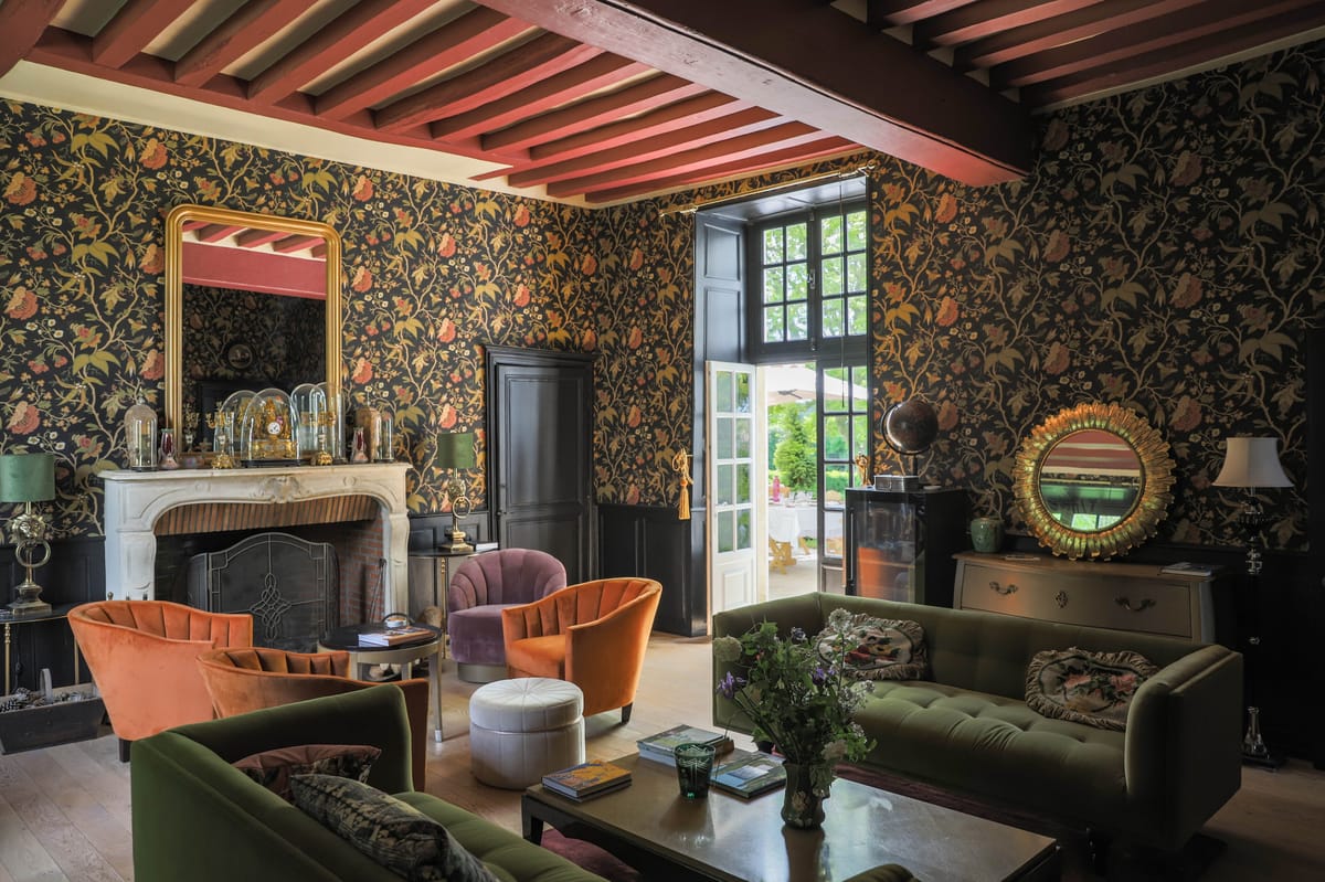 Maximalist salon with dark floral wallpaper, green velvet sofas, burnt orange chairs, and marble fireplace