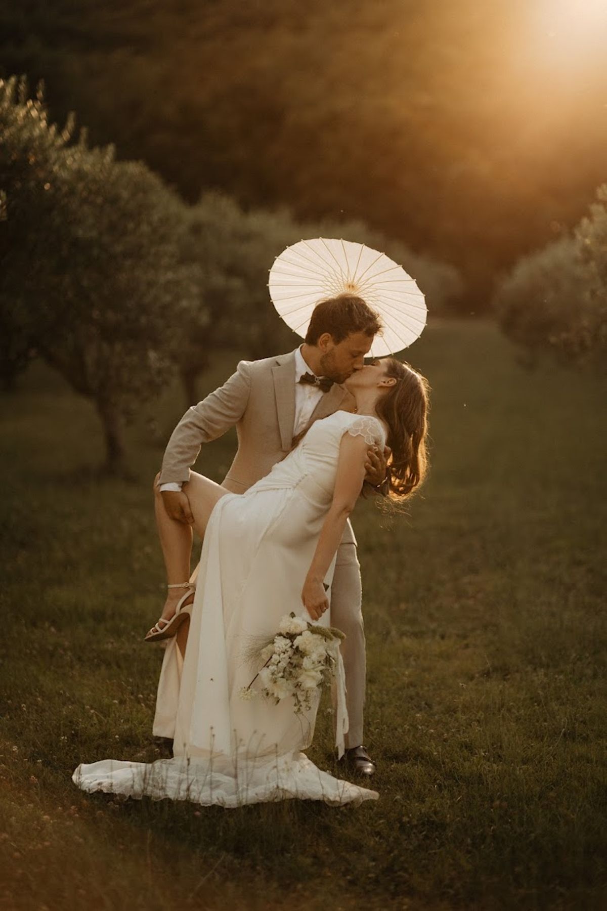 Dip kiss in olive grove at golden hour, groom in sand linen suit, bride with ranunculus bouquet and parasol