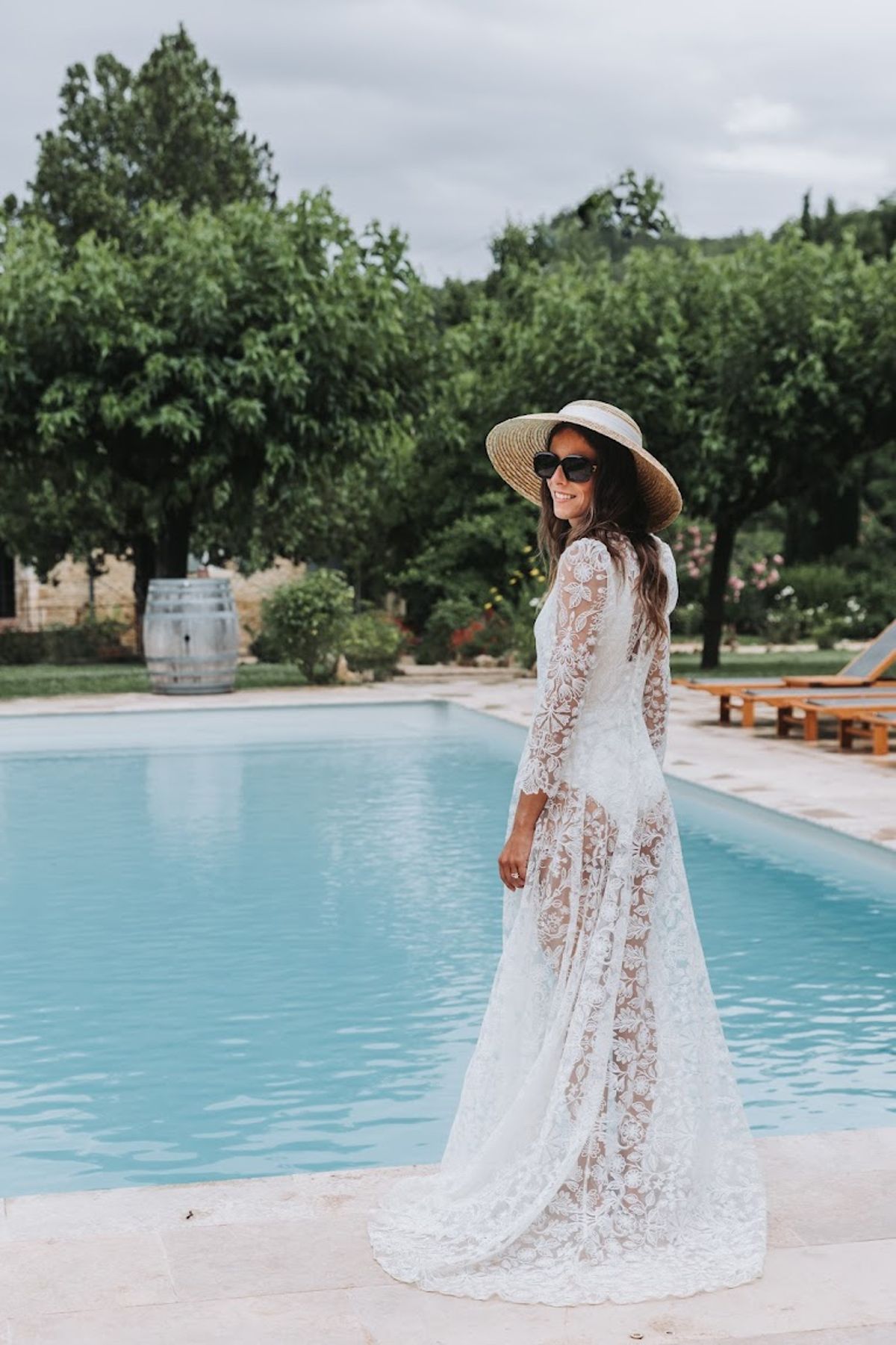 Bride in sheer lace gown with straw boater hat and black sunglasses posing beside swimming pool at estate