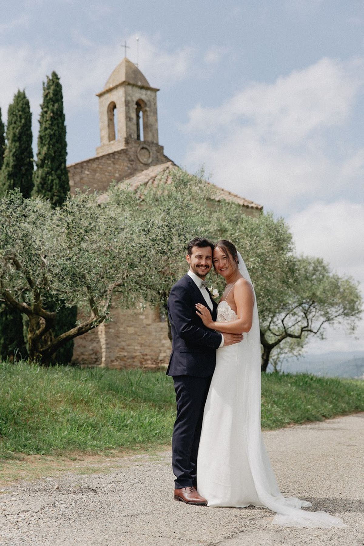 Couple embracing before Romanesque stone chapel with bell tower flanked by cypress and olive trees