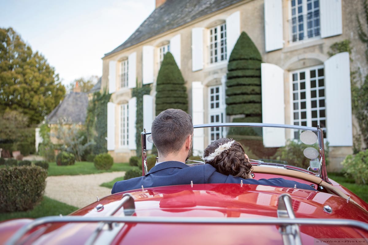 Couple seated in red vintage convertible facing a French manor house with stone facade and white shutters