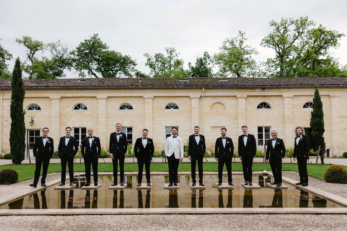 Groom and eleven groomsmen in formal tuxedos standing by reflecting pool at classical stone venue