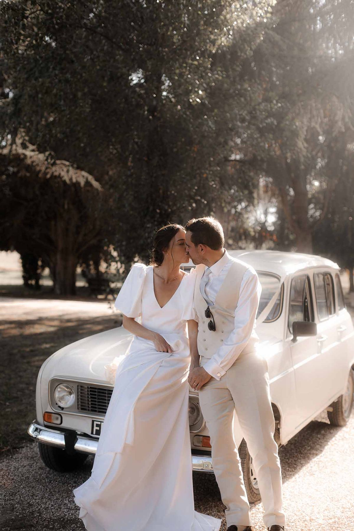 Bride and groom posing on vintage white car on tree-lined country road during wedding portrait session