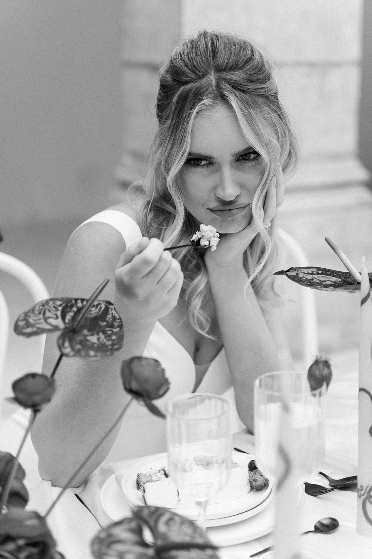 Bride playfully offers fork of wedding cake to camera at editorial reception table in B&W