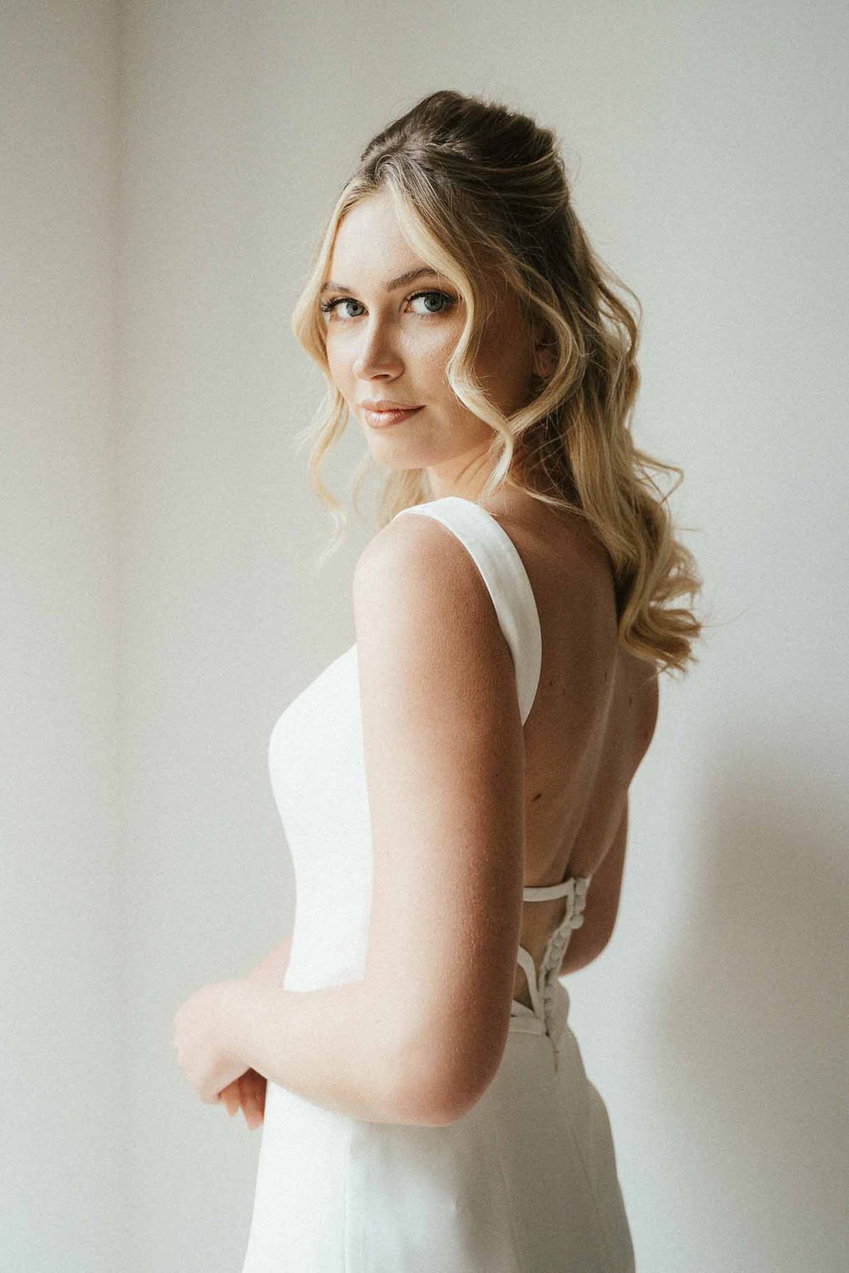 Rear three-quarter portrait of bride in open-back sleeveless gown with soft waves against white wall