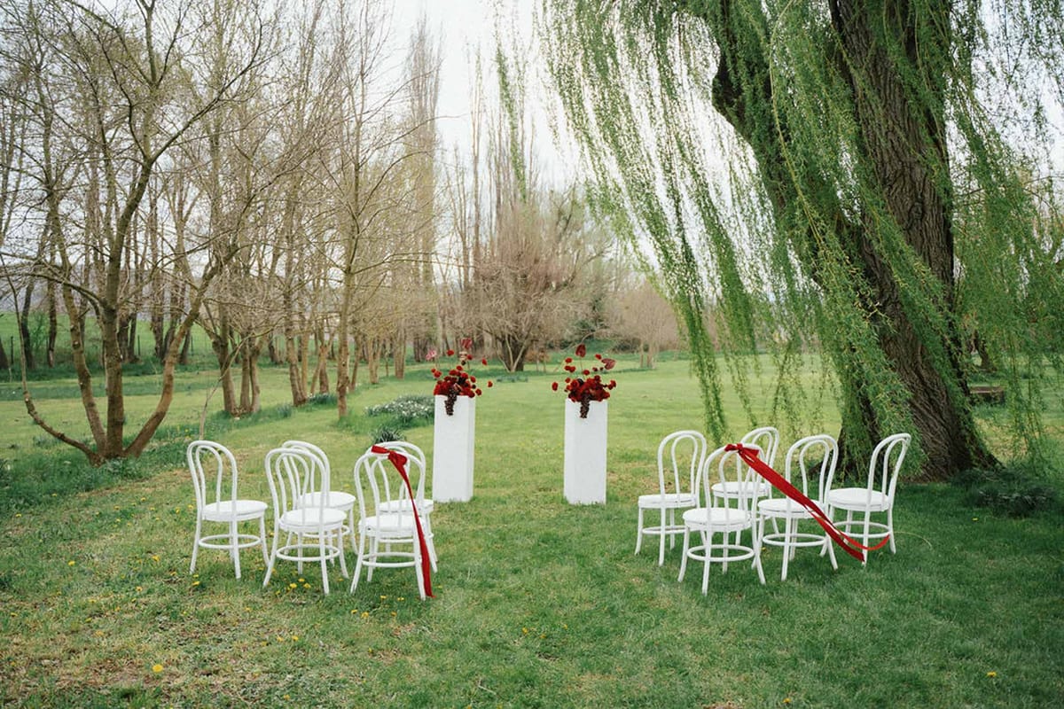 White bentwood chairs with deep red satin ribbons and red anthurium pedestal arrangements beside willow tree