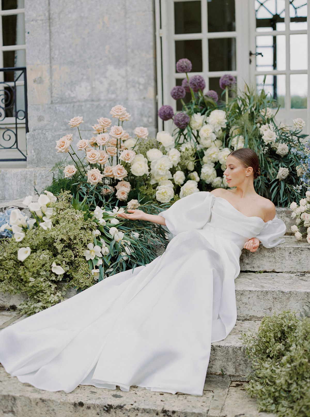 Bride in off-shoulder white gown poses in stone courtyard with elaborate floral installations