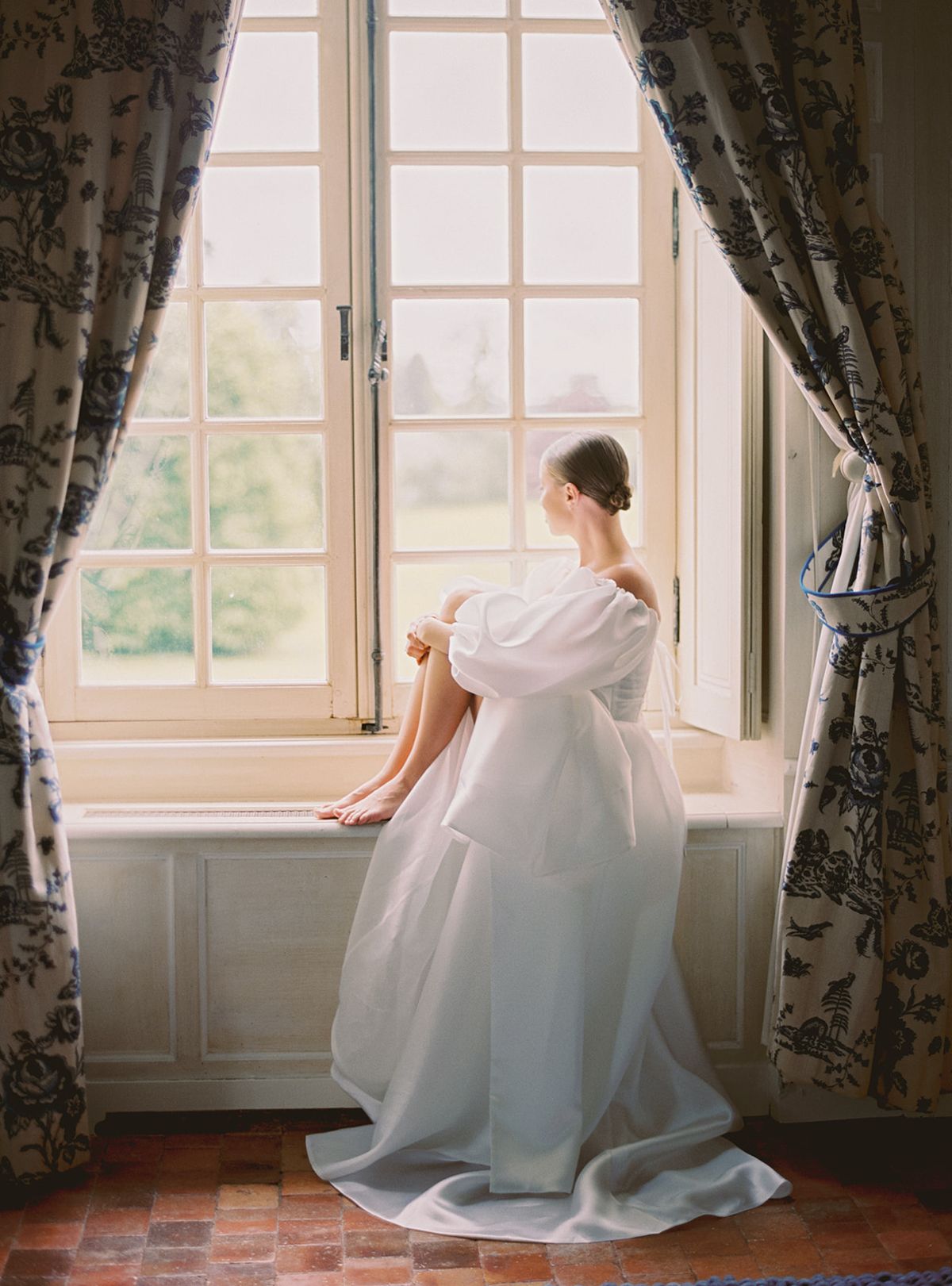 Bride in flowing white gown sitting by window in elegant period room with botanical curtains