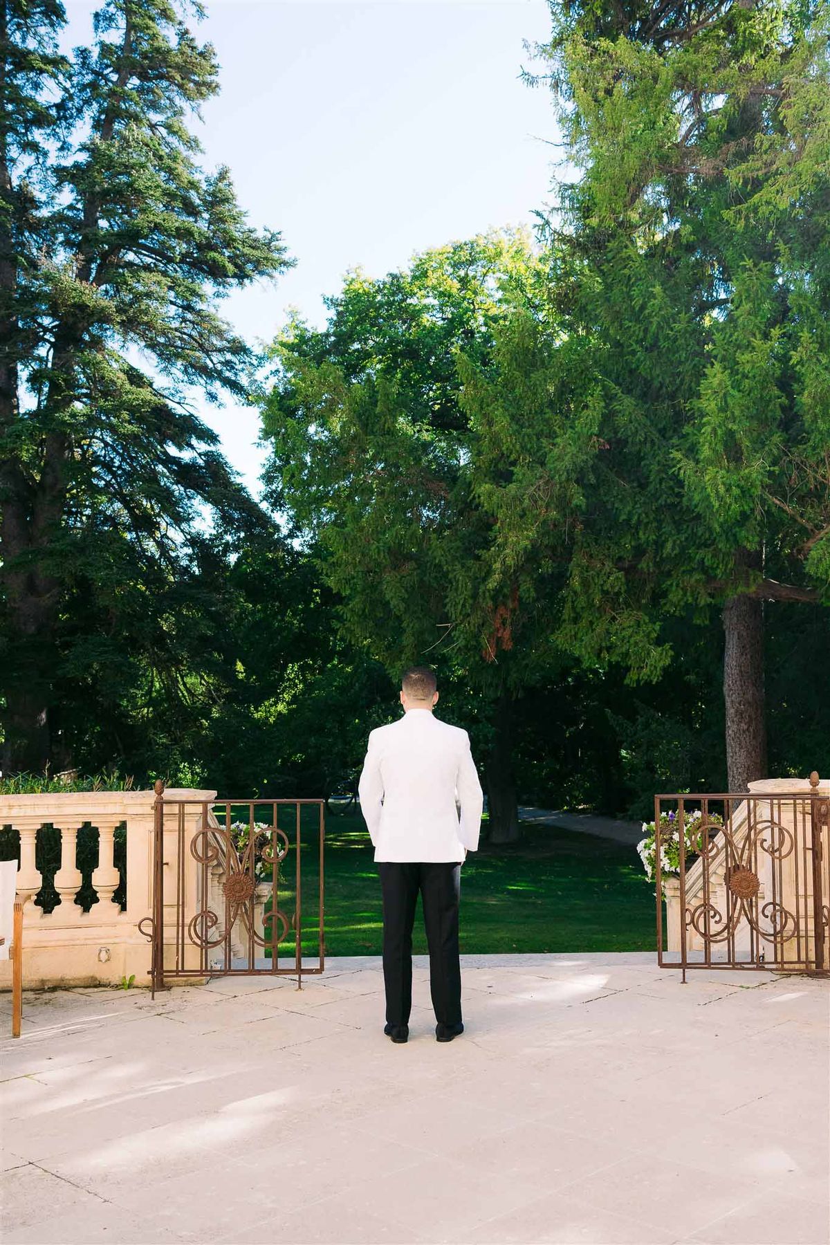 Groom in white jacket standing before ornamental gates at formal garden venue