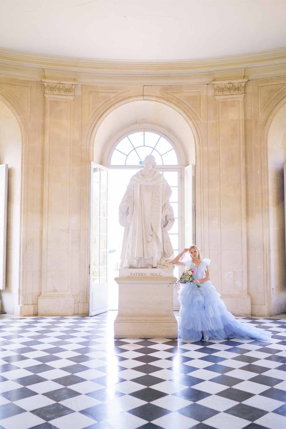 Bride in blue tulle gown with bouquet poses beside marble statue in classical palace interior