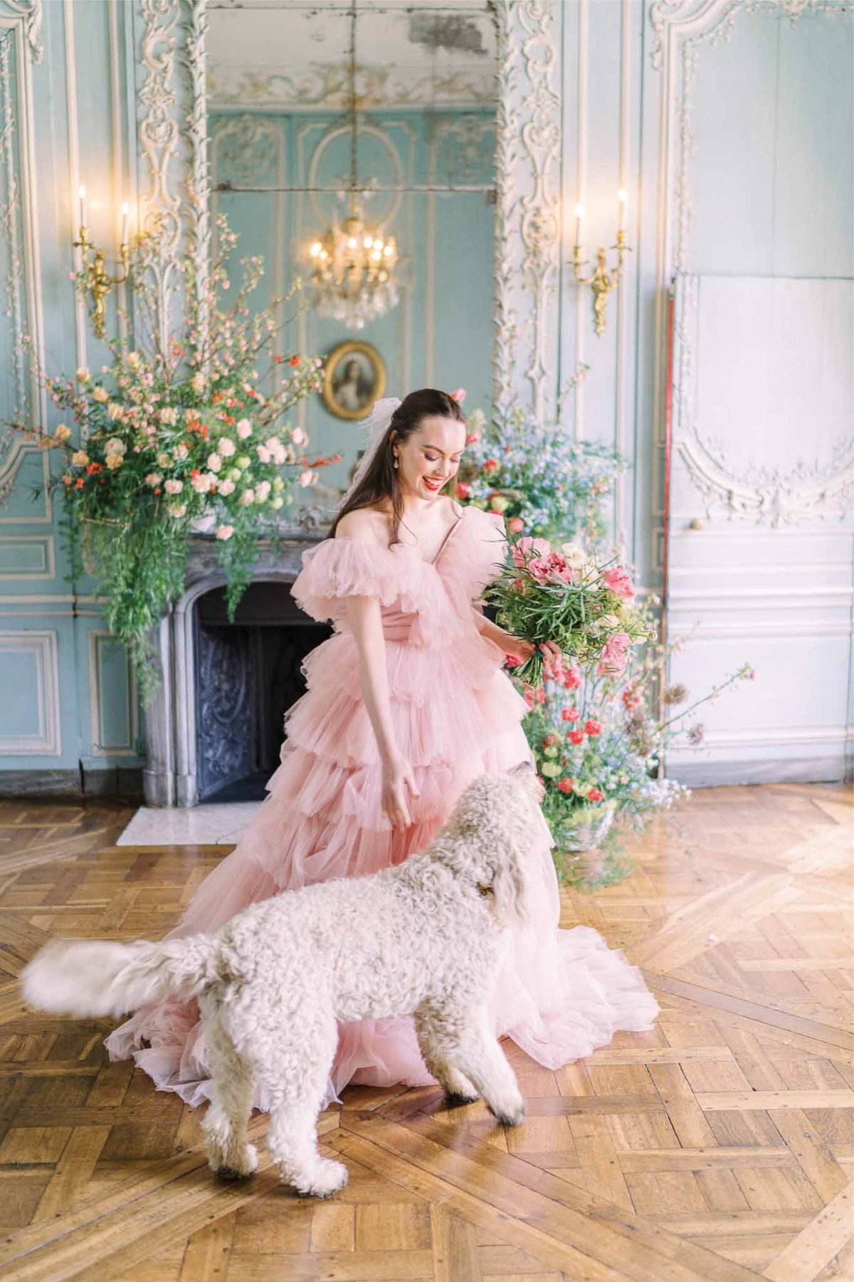 Bride in blush pink gown with bouquet and white dog in neoclassical ballroom with crystal chandelier