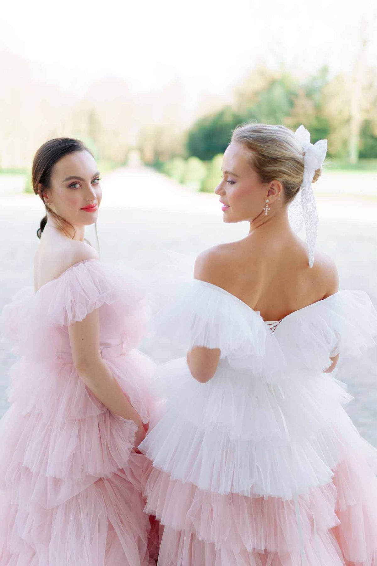 Two women in ivory and blush tulle dresses posing along tree-lined garden path