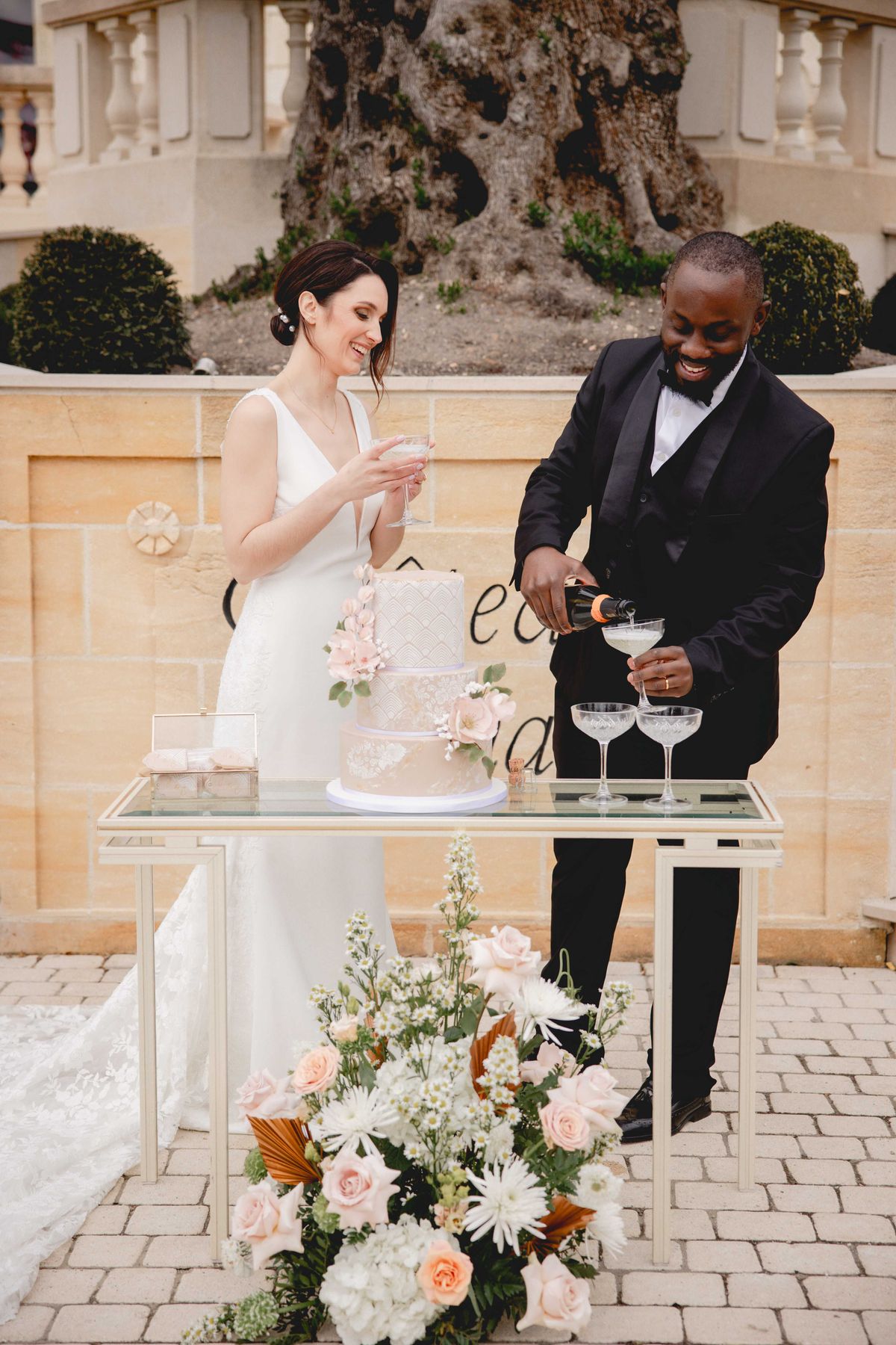 Bride and groom pouring champagne at gold-framed cake table on cobblestone terrace with three-tier blush wedding cake