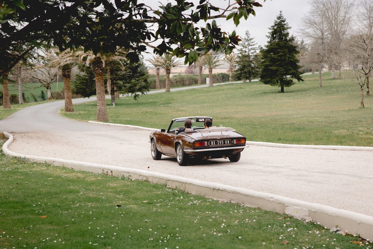 Couple driven away in vintage brown convertible along curved gravel driveway lined with palm trees at estate