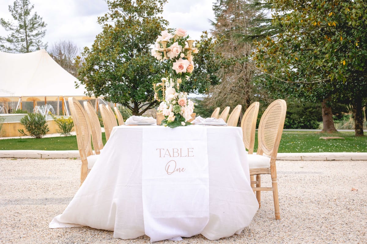 Round table with blush rose candelabra centerpiece, cane-back chairs, and printed table runner on gravel