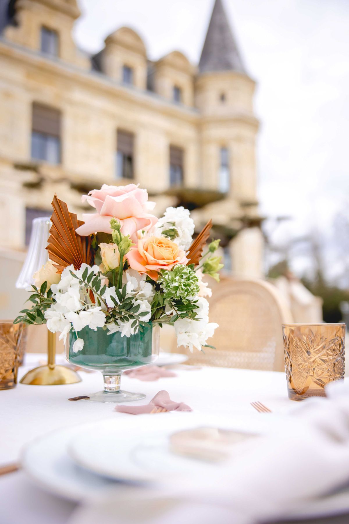 Teal compote vase with peach roses and dried palm leaves beside amber glasses and gold flatware