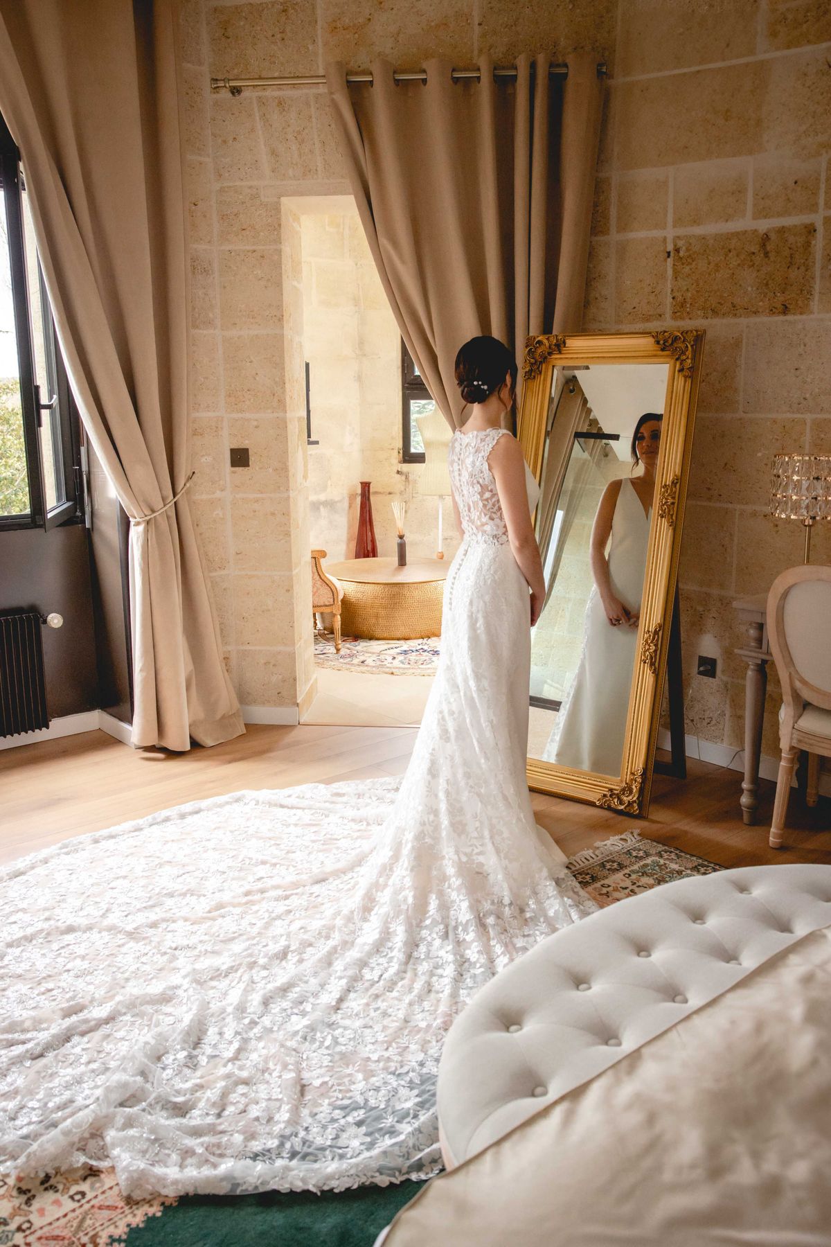 Bride from behind in scoop-back lace gown with cathedral train viewing reflection in gold mirror in limestone bedroom
