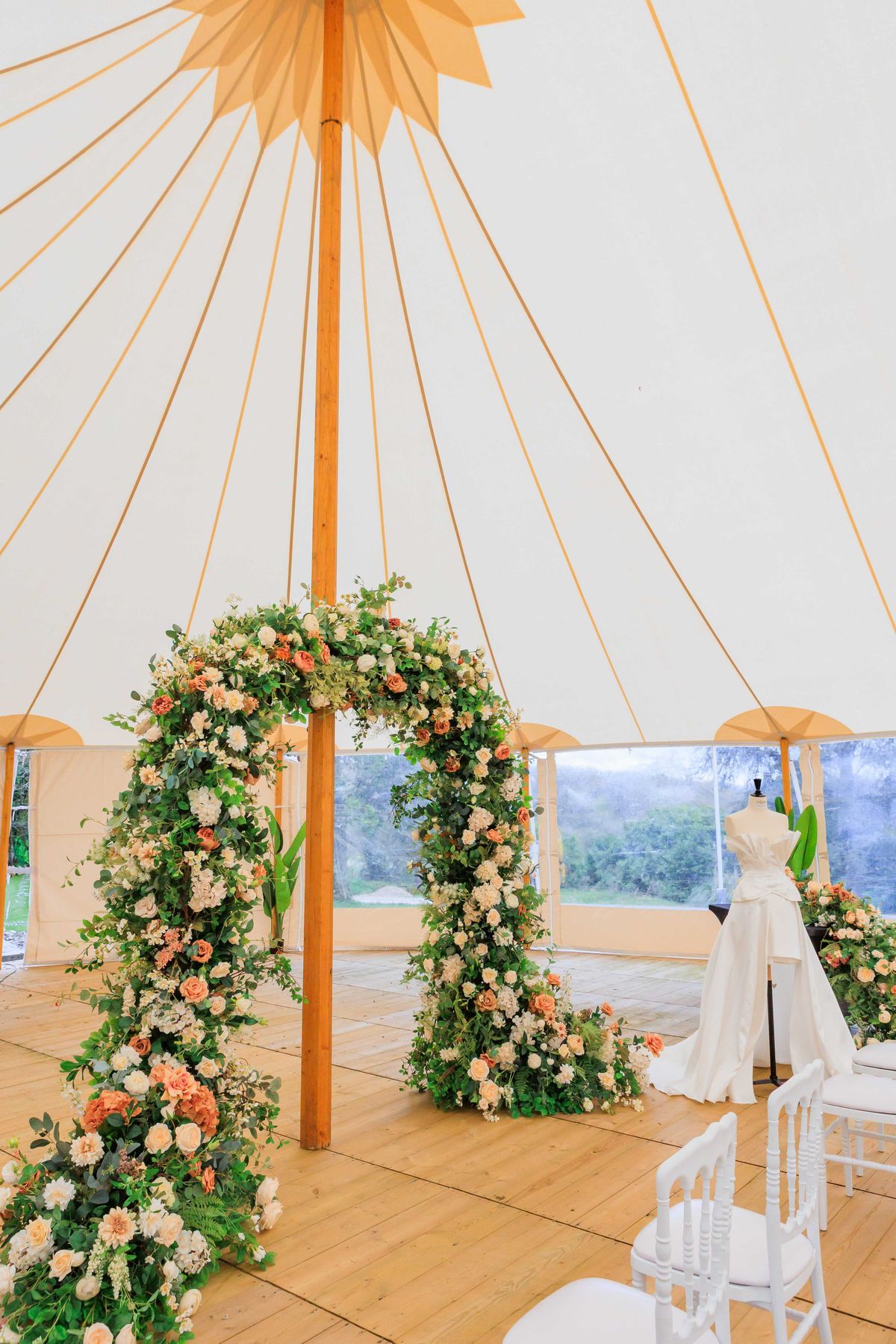 Sperry tent ceremony with circular floral arch of peach roses and ivory dahlias beside displayed gown