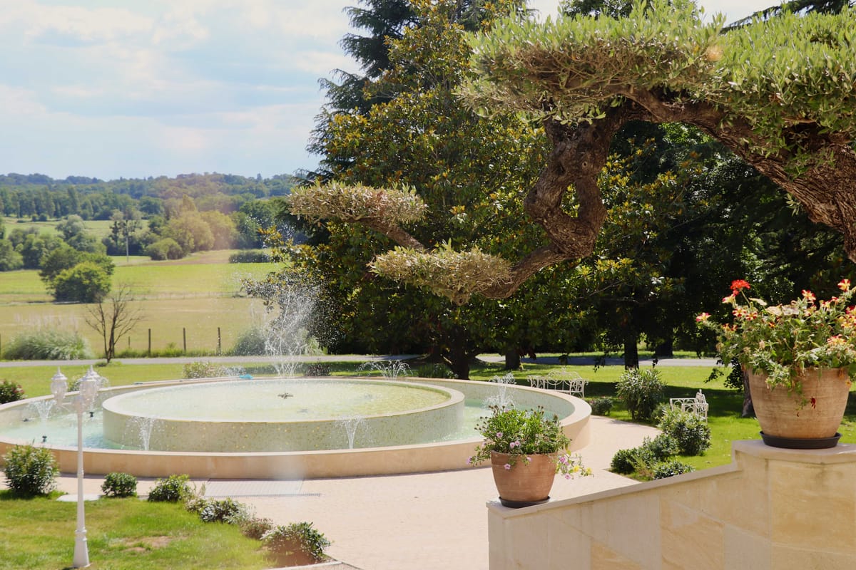 Circular stone fountain with water jets on cream terrace with terracotta planters and countryside view