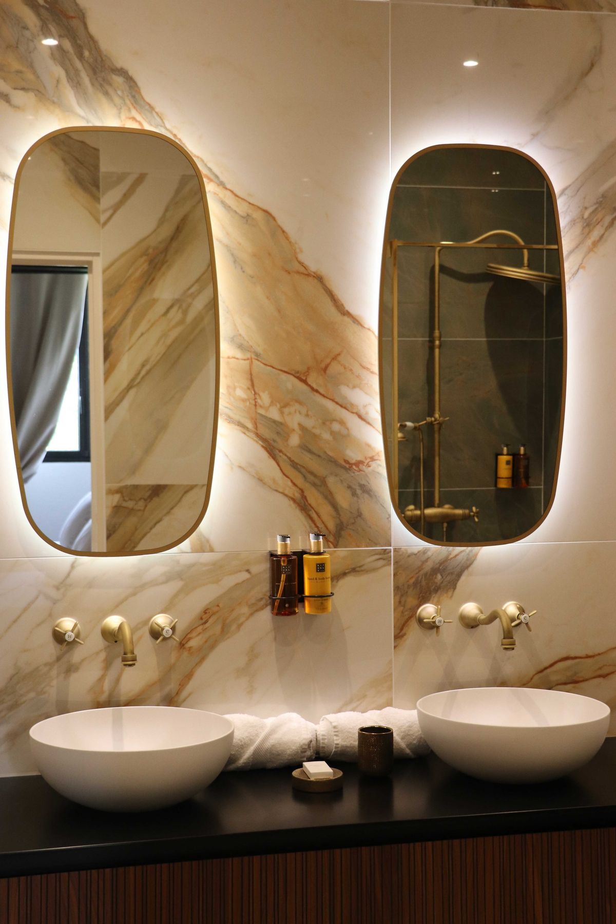 Luxury bathroom with white vessel sinks, amber marble wall, brushed brass fixtures, and backlit oval mirrors